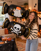 Party Paradise Store woman holding two black candy-shaped Halloween balloons featuring ghost and skull designs.